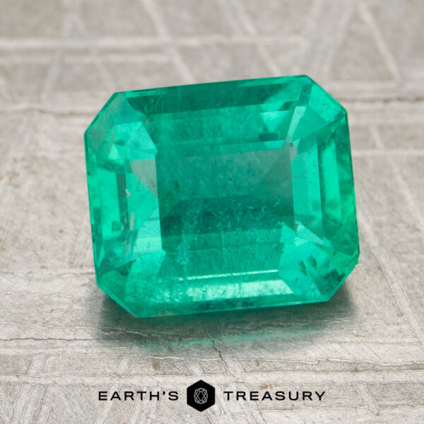 2.45-Carat Bright Green Brazilian Emerald A 2.45-carat bright green Brazilian emerald with an emerald cut sits on a textured surface. The words "EARTH'S TREASURY" and a hexagon logo appear at the bottom of the image.