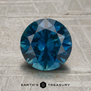 A 2.24-carat rich peacock blue Montana sapphire (heated), cut in a round brilliant style, displayed on a textured surface with the Earth's Treasury logo and text at the bottom.