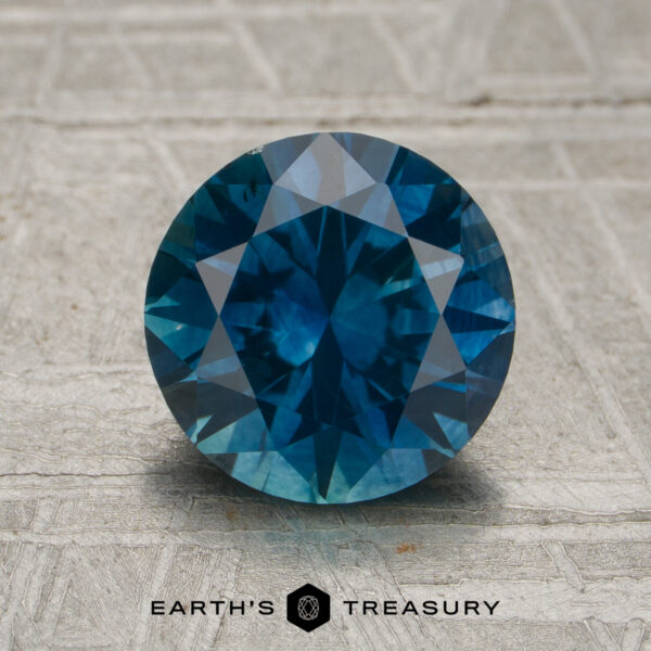 2.24-Carat Rich Peacock Blue Montana Sapphire (Heated) A 2.24-carat rich peacock blue Montana sapphire (heated), cut in a round brilliant style, displayed on a textured surface with the Earth's Treasury logo and text at the bottom.