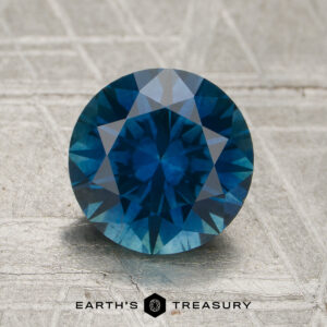 A 2.21-carat rich peacock blue Montana sapphire (heated) in a round brilliant cut is displayed on a textured surface with the "Earth's Treasury" logo and text at the bottom of the image.