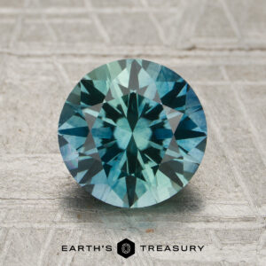 2.17-carat aqua blue-green particolored Montana sapphire (heated), round brilliant-cut, displayed on a textured gray background with the "Earth's Treasury" logo at the bottom.