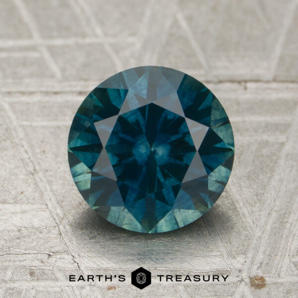 A 2.17-carat Peacock Montana Sapphire (heated) in a round cut is displayed on a textured, light-colored surface with the "Earth's Treasury" logo and text beneath it.