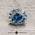 A 1.39-carat teal to blue Montana sapphire (heated), faceted in an oval shape, rests on a textured gray surface, with the words "EARTH'S TREASURY" and a hexagon logo below it.