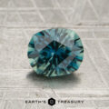 A 1.39-carat teal to blue Montana sapphire (heated) is displayed on a textured gray surface with the text "EARTH'S TREASURY" and a small logo beneath it.