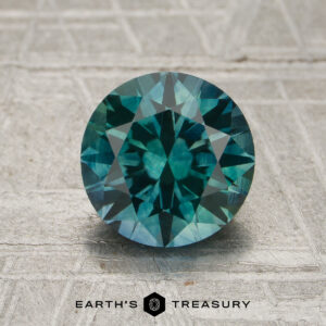 1.92-carat round brilliant-cut spruce green Montana sapphire (heated) showcased on a textured surface, featuring "Earth's Treasury" branding at the bottom.