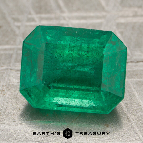 3.43-Carat Rich Green Zambian Emerald A 3.43-carat rich green Zambian emerald, cut in a rectangular faceted shape, rests on a textured surface above the "EARTH'S TREASURY" name and geometric logo.