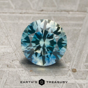 A 1.81-carat blue-aqua particolored Montana sapphire (heated) is showcased on a textured gray background, with the "EARTH'S TREASURY" logo and text at the bottom.