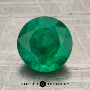 A 2.34-carat rich green Zambian emerald, expertly round-cut, is displayed on a textured gray surface with the "Earth's Treasury" logo and text beneath it.
