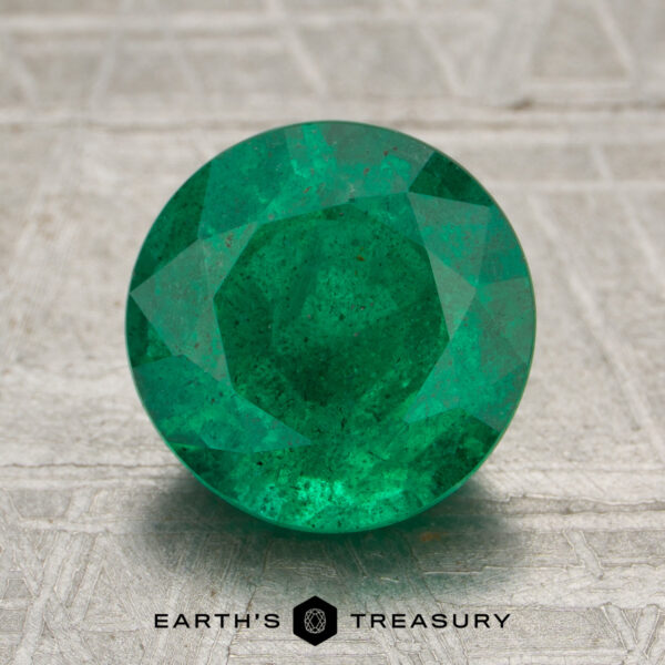 2.34-Carat Rich Green Zambian Emerald A 2.34-carat rich green Zambian emerald, expertly round-cut, is displayed on a textured gray surface with the "Earth's Treasury" logo and text beneath it.