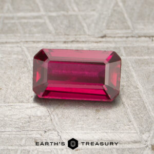 A rectangular, faceted 1.02-carat Mozambique ruby (heated) is displayed on a textured gray surface, with the text "EARTH'S TREASURY" at the bottom of the image.