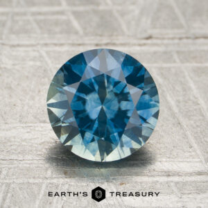 1.82-carat Montana teal to violet blue color-change sapphire, round brilliant-cut, displayed on a textured surface with the "Earth's Treasury" logo and text at the bottom.