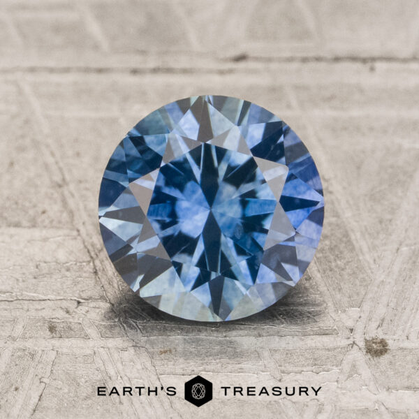 1.71-Carat Teal to Violet Blue Color-Change Montana Sapphire (Heated) Certainly! Here is the rewritten sentence using the provided product data: A 1.71-carat teal to violet blue color-change Montana sapphire in a round brilliant cut is displayed on a textured gray surface, with the "Earth's Treasury" text and logo positioned beneath.