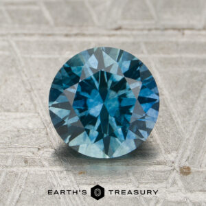 A 1.71-carat teal to violet blue color-change Montana sapphire, cut in a round brilliant style, is displayed on a textured surface. The Earth's Treasury logo and name appear at the bottom of the image.