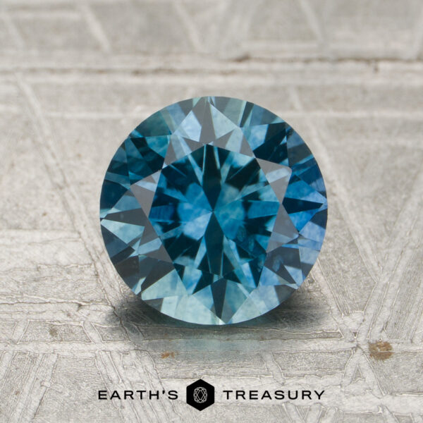 1.71-Carat Teal to Violet Blue Color-Change Montana Sapphire (Heated) A 1.71-carat teal to violet blue color-change Montana sapphire, cut in a round brilliant style, is displayed on a textured surface. The Earth's Treasury logo and name appear at the bottom of the image.