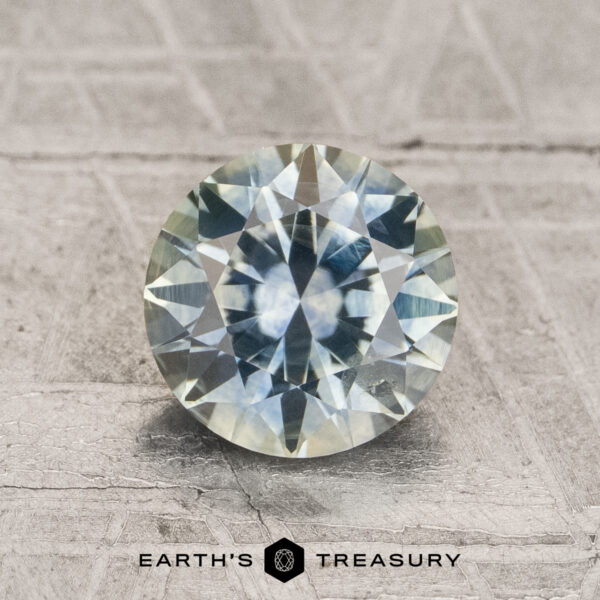 1.66-Carat Mint Green to Gray Color-Change Montana Sapphire (Heated) A round, faceted 1.66-carat Montana sapphire, exhibiting aqua to steely blue-gray color-change hues, is displayed on a textured surface. "EARTH'S TREASURY" is written at the bottom of the image.