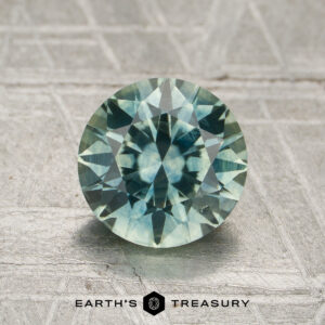 A 1.66-carat aqua to steely blue-gray color-change Montana sapphire, cut into a round, faceted shape, is displayed upright on a textured surface with the words "Earth's Treasury" below it.