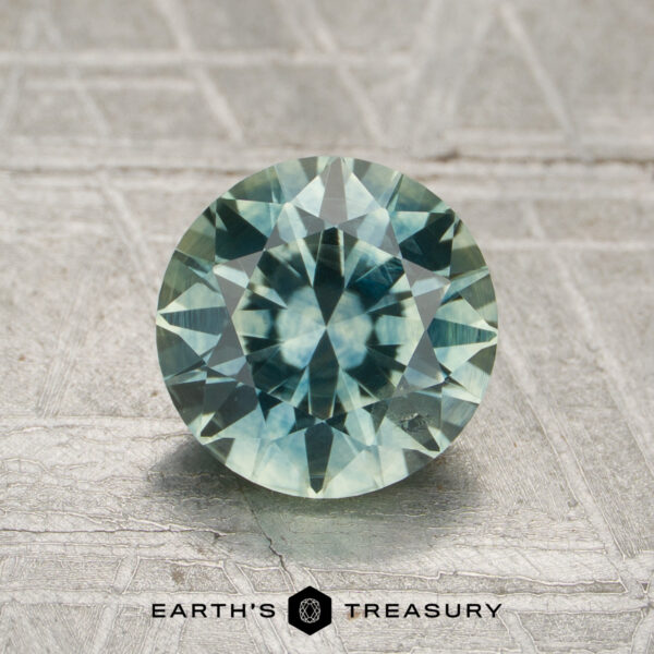 1.66-Carat Mint Green to Gray Color-Change Montana Sapphire (Heated) A 1.66-carat aqua to steely blue-gray color-change Montana sapphire, cut into a round, faceted shape, is displayed upright on a textured surface with the words "Earth's Treasury" below it.