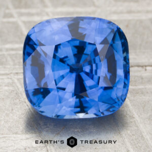 A 4.56-carat cornflower blue Ceylon sapphire (heated), cushion-cut, is displayed on a textured surface featuring the words "EARTH'S TREASURY" and a hexagonal logo at the bottom.
