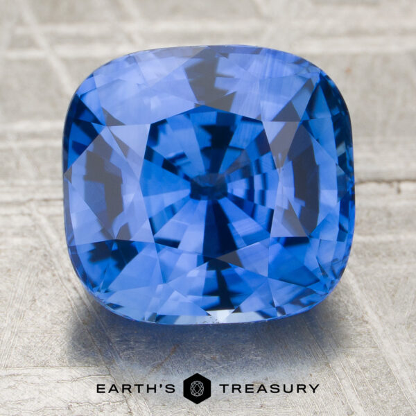 4.56-Carat Cornflower Blue Ceylon Sapphire (Heated) A 4.56-carat cornflower blue Ceylon sapphire (heated), cushion-cut, is displayed on a textured surface featuring the words "EARTH'S TREASURY" and a hexagonal logo at the bottom.