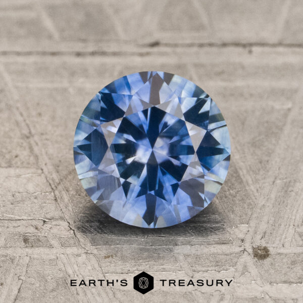 1.61-Carat Teal Blue to Violet Color-Change Montana Sapphire (Heated) A 1.61-carat teal blue to purple color-change Montana sapphire (Heated) in a round brilliant cut displayed on a textured surface, with the "Earth's Treasury" logo and text at the bottom of the image.