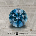 A 1.61-carat, round brilliant-cut Montana sapphire—exhibiting a striking color change from teal blue to purple—is displayed upright on a textured gray surface, with the text "EARTH'S TREASURY" beneath it.