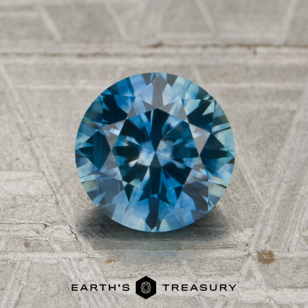 1.61-Carat Teal Blue to Violet Color-Change Montana Sapphire (Heated) A 1.61-carat, round brilliant-cut Montana sapphire—exhibiting a striking color change from teal blue to purple—is displayed upright on a textured gray surface, with the text "EARTH'S TREASURY" beneath it.