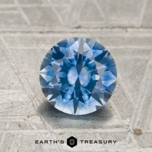 A round, faceted 1.55-carat medium blue to purple color-change Montana sapphire is displayed on a textured silver surface, with the "Earth's Treasury" logo and text at the bottom.
