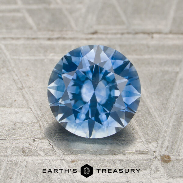 1.55-Carat Sky Blue to Purple Color-Change Montana Sapphire (Heated) A round, faceted 1.55-carat medium blue to purple color-change Montana sapphire is displayed on a textured silver surface, with the "Earth's Treasury" logo and text at the bottom.
