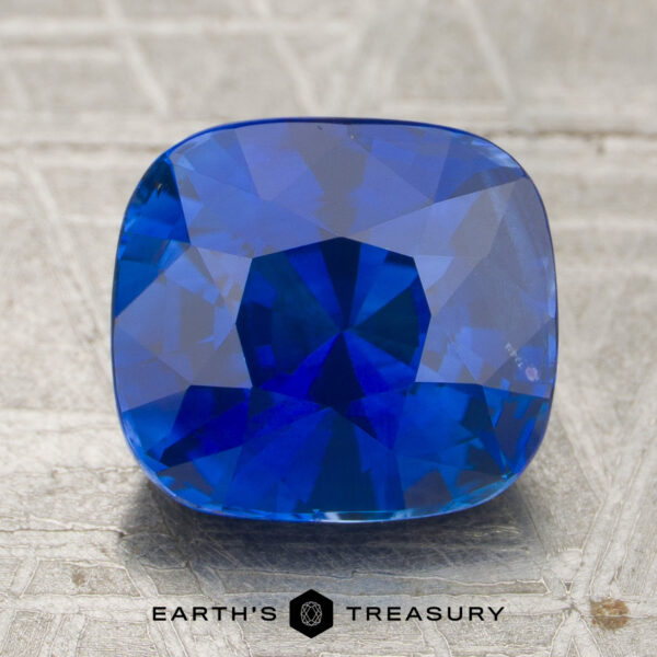 3.45-Carat Royal Blue Ceylon Sapphire (Heated) A 3.45-carat royal blue Ceylon sapphire (heated), expertly cut in a cushion shape, is displayed on a textured surface with the Earth’s Treasury logo visible at the bottom of the image.