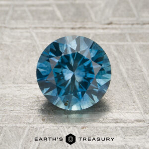 A 1.49-carat teal blue to violet color-change Montana sapphire with faceted cuts displayed on a textured light background, featuring the "Earth's Treasury" logo and text at the bottom.