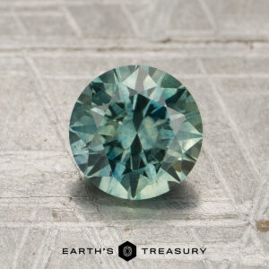 A 1.45-carat round-cut heated Montana sapphire in blue-green hues displayed on a textured gray surface with the words "Earth's Treasury" below the gemstone.