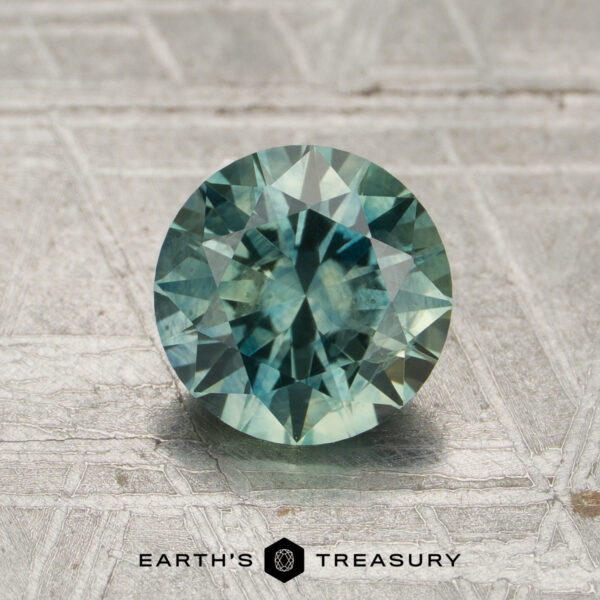 1.45-Carat Spruce Green Montana Sapphire (Heated) A 1.45-carat round-cut heated Montana sapphire in blue-green hues displayed on a textured gray surface with the words "Earth's Treasury" below the gemstone.