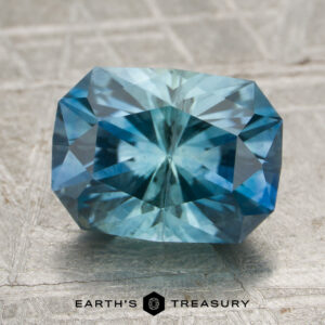 A 2.57-carat aqua-blue particolored Montana sapphire (heated), featuring a rectangular cushion cut, rests on a textured surface with the “Earth’s Treasury” logo and text beneath it.