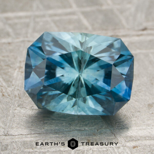 2.57-Carat Aqua-Blue Particolored Montana Sapphire (Heated) A 2.57-carat aqua-blue particolored Montana sapphire (heated), featuring a rectangular cushion cut, rests on a textured surface with the “Earth’s Treasury” logo and text beneath it.