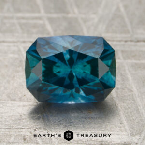 A 2.01-carat rich blue-teal particolored Montana sapphire (heated), cut in a rectangular shape with multiple facets, is displayed on a textured surface. "EARTH'S TREASURY" appears at the bottom along with a hexagonal logo.