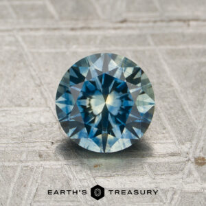 A 1.30-carat blue-yellow to violet-yellow particolored color-change gemstone, expertly round brilliant-cut, is displayed upright on a textured metallic surface with "Earth's Treasury" text at the bottom.