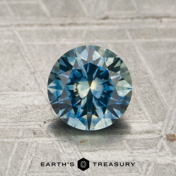 1.30-Carat Teal-Yellow Particolored Montana Sapphire (Heated) A 1.30-carat blue-yellow to violet-yellow particolored color-change gemstone, expertly round brilliant-cut, is displayed upright on a textured metallic surface with "Earth's Treasury" text at the bottom.