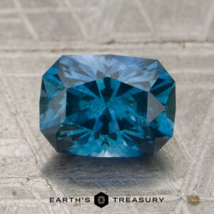 A rectangular, rich teal blue 1.99-carat Montana sapphire (heated) with faceted cuts rests on a textured, metallic surface featuring the "EARTH'S TREASURY" text and logo below.