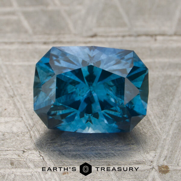 1.99-Carat Rich Teal Blue Montana Sapphire (Heated) A rectangular, rich teal blue 1.99-carat Montana sapphire (heated) with faceted cuts rests on a textured, metallic surface featuring the "EARTH'S TREASURY" text and logo below.