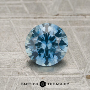 A 1.11-carat sky blue to violet color-change Montana sapphire is placed on a textured grey surface, with the “EARTH’S TREASURY” text and a hexagon logo visible at the bottom.