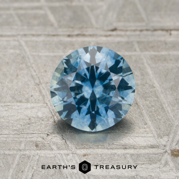 1.11-Carat Sky Blue Montana Sapphire (Heated) A 1.11-carat sky blue to violet color-change Montana sapphire is placed on a textured grey surface, with the “EARTH’S TREASURY” text and a hexagon logo visible at the bottom.
