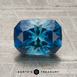 A 1.91-carat rich blue-teal particolored Montana sapphire (heated) with intricate facets is displayed on a textured surface, with the Earth's Treasury logo and text below.