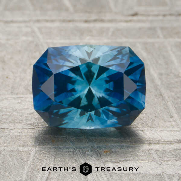 1.91-Carat Rich Blue-Teal Particolored Montana Sapphire (Heated) A 1.91-carat rich blue-teal particolored Montana sapphire (heated) with intricate facets is displayed on a textured surface, with the Earth's Treasury logo and text below.