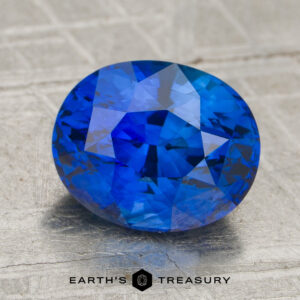 A 2.63-carat royal blue Ceylon sapphire (heated) in an oval faceted cut rests on a textured gray surface, with the "Earth's Treasury" logo and text displayed beneath it.