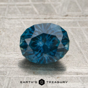 A 1.77-carat peacock blue Montana sapphire (heated) with an oval cut is displayed on a textured gray surface. The "Earth's Treasury" logo and text appear at the bottom of the image.