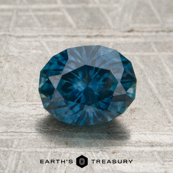 A 1.77-carat peacock blue Montana sapphire (heated) with an oval cut is displayed on a textured gray surface. The "Earth's Treasury" logo and text appear at the bottom of the image.