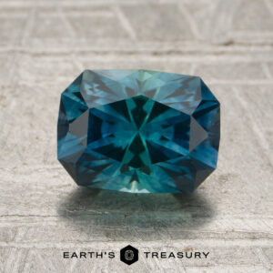 A 1.74-carat peacock blue-teal particolored Montana sapphire (heated), expertly faceted in a rectangular shape, rests on a textured gray surface with the “Earth’s Treasury” logo and text displayed at the bottom.