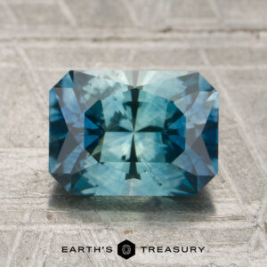 A 1.87-carat aqua-blue particolored Montana sapphire, featuring a rectangular, faceted cut, is displayed on a textured surface with the "Earth's Treasury" logo beneath it.