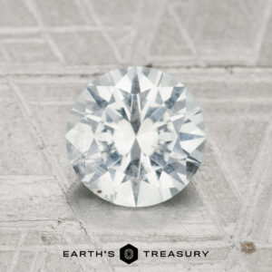 A 1.58-carat Montana sapphire is displayed on a textured gray surface, with "EARTH'S TREASURY" and a geometric logo visible at the bottom.