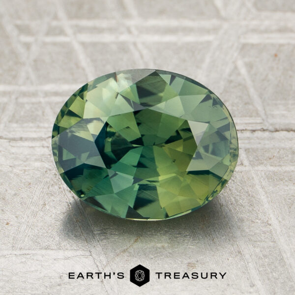 2.34-Carat Yellowish Green Madagascar Sapphire (Heated) A 2.34-carat yellowish green Madagascar sapphire (heated), cut in an oval shape, rests on a textured gray surface with the words "Earth's Treasury" below it.